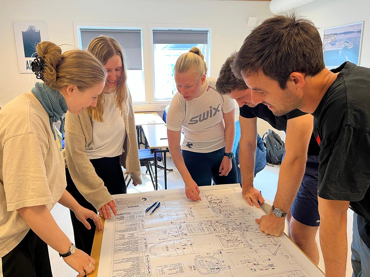 Redningsdrakt-kurs.jpg Flere personer står rundt et bord og diskuterer.